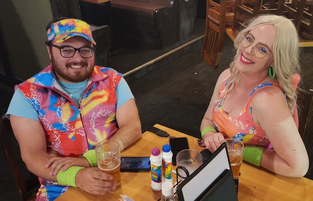 Two players in colourful 80s outfits smile at a pub table with dabber markers and drinks